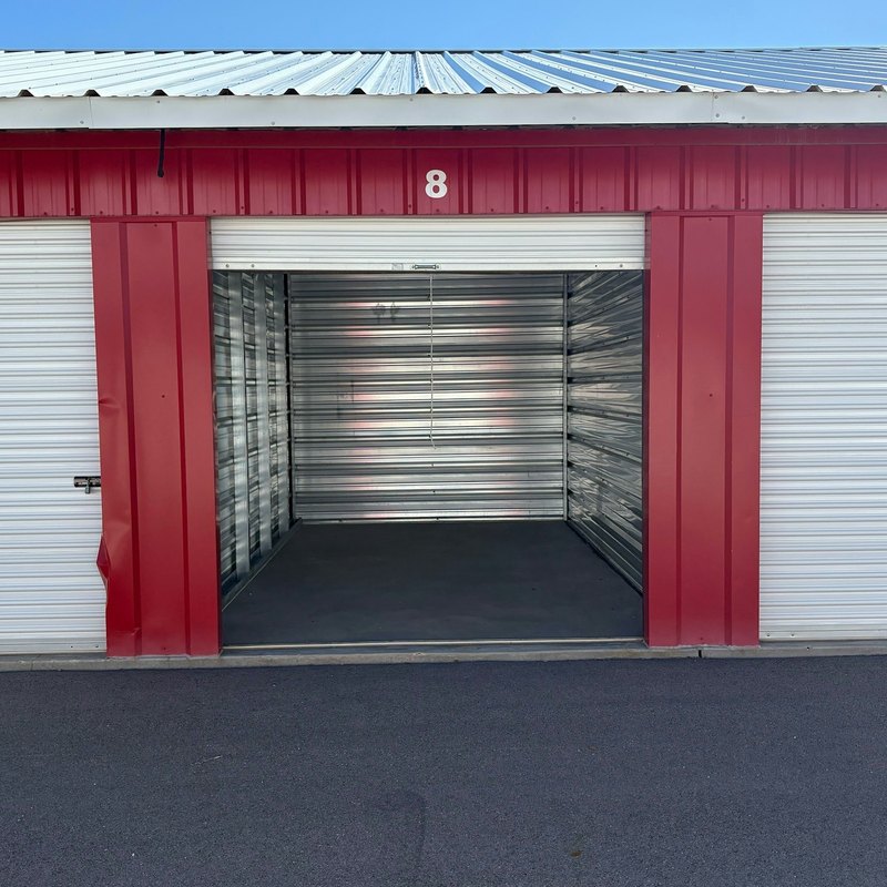 Storage unit with its white roll-up door raised on an empty unit with metal walls, between red door frames under a metal roof
