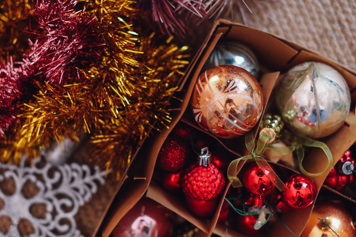Glass Christmas ornaments in gold, silver and red packed in a divided cardboard box beside tinsel garland