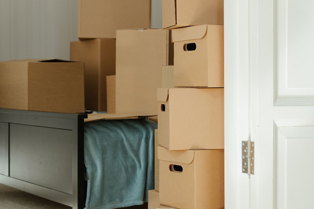 Stacked cardboard moving boxes and a bed frame in a bedroom seen through an open white door