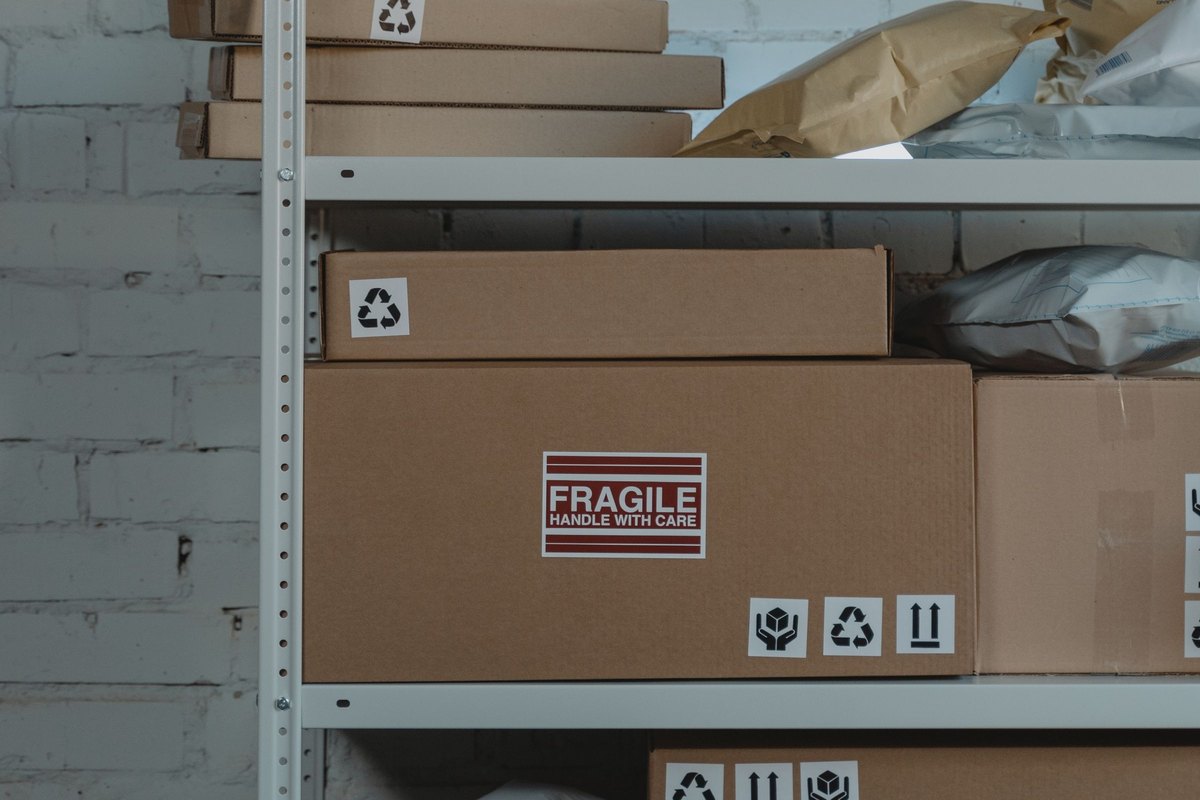 Metal shelving against a white brick wall holding cardboard boxes, one with a red Fragile label, and padded mailers