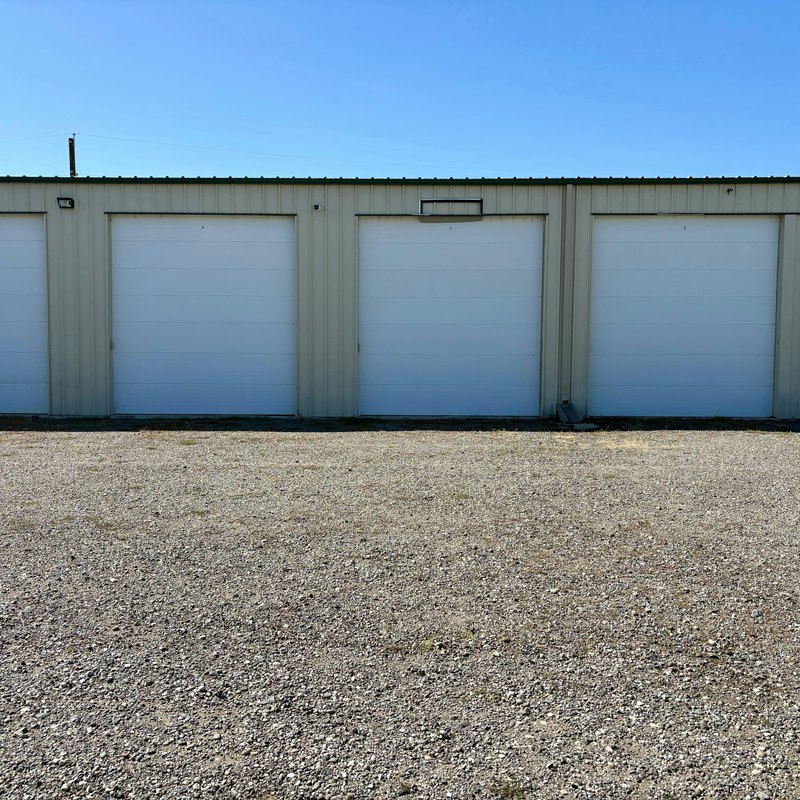 Row of white roll-up storage doors on a beige metal building above a gravel lot